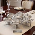 Paramount Series 7-in-1 buffet food warmer set featuring seven clear glass bowls with polished stainless steel lids and a silver metal stand with buffet burners on a decorated dining table.
