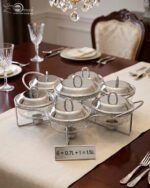 Paramount Series 7-in-1 buffet food warmer set featuring seven clear glass bowls with polished stainless steel lids and a silver metal stand with buffet burners on a decorated dining table.