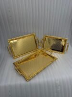 A luxury three-piece set of rectangular gold-plated stainless steel serving trays with intricate floral embossing and ornate vintage handles, displayed on a wooden dining table with elegant place settings.