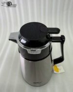 A 1-liter stainless steel vacuum thermal flask with a black insulated lid and ergonomic handle, placed on a kitchen countertop next to lemons and a coffee mug.