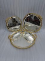 Set of three round silver stainless steel serving trays with floral etched patterns, gold rope borders, and ornate gold handles on a wooden dining table.