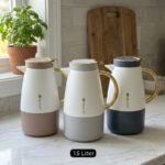 Three elegant 1.5-liter stainless steel thermal flasks in rose gold, slate grey, and charcoal blue bases with white bodies and gold-plated handles, displayed on a marble kitchen countertop.