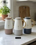 Three elegant 1.5-liter stainless steel thermal flasks in rose gold, slate grey, and charcoal blue bases with white bodies and gold-plated handles, displayed on a marble kitchen countertop.
