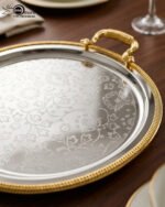 Set of three round silver stainless steel serving trays with floral etched patterns, gold rope borders, and ornate gold handles on a wooden dining table.