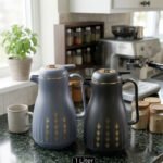Two elegant 1-liter thermal carafes in matte black and slate blue with gold decorative patterns, displayed on a kitchen counter next to a coffee machine.