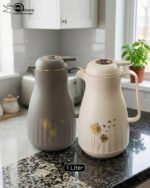Two 1-liter insulated thermal flasks in Earth Grey and Creamy Beige, featuring golden dandelion designs and ergonomic handles, displayed on a kitchen countertop.