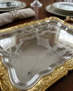 A three-piece set of rectangular stainless steel serving trays featuring polished silver mirror bases with etched ribbon designs and ornate gold-embossed scalloped borders, displayed on a dark wooden dining table.