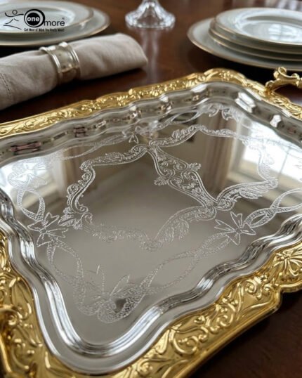 A three-piece set of rectangular stainless steel serving trays featuring polished silver mirror bases with etched ribbon designs and ornate gold-embossed scalloped borders, displayed on a dark wooden dining table.