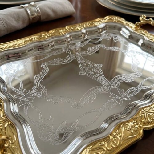 A three-piece set of rectangular stainless steel serving trays featuring polished silver mirror bases with etched ribbon designs and ornate gold-embossed scalloped borders, displayed on a dark wooden dining table.
