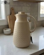 A 1-liter insulated thermal flask with a 3D diamond-patterned matte body, featuring a curved handle and a spout, shown in black and beige color options on a kitchen countertop.