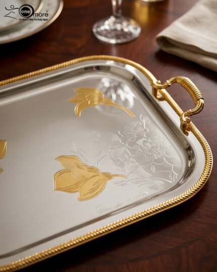 A set of three nesting stainless steel rectangular serving trays with gold rope borders, floral engravings, and ornate gold handles, displayed on a dark wooden dining table with elegant dinnerware.