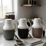 Three 1-liter Feenik thermal flasks in charcoal, taupe, and gray colors with gold trim on a marble kitchen countertop.