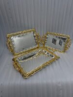 A three-piece set of rectangular stainless steel serving trays featuring polished silver mirror bases with etched ribbon designs and ornate gold-embossed scalloped borders, displayed on a dark wooden dining table.