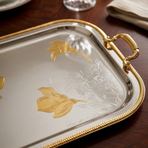 A set of three nesting stainless steel rectangular serving trays with gold rope borders, floral engravings, and ornate gold handles, displayed on a dark wooden dining table with elegant dinnerware.