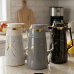 Three elegant 1-liter vacuum flasks in black, grey, and cream colors, featuring a golden Kintsugi-inspired marble pattern on a kitchen countertop.