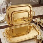 Three-piece set of luxury gold-plated stainless steel serving trays with intricate floral engravings and textured handles, displayed on a formal dining table.