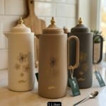 Three 1-liter DayDays insulated flasks in white, taupe, and grey, featuring gold floral designs and decorative gold lids on a kitchen counter.