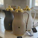 Three 1-liter thermal flasks in grey, beige, and white featuring matte finishes, elegant gold lids, and ergonomic handles on a marble countertop.