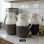 Three 1-liter modern vacuum flasks with cream-colored tops and ribbed bases in brown, grey, and beige, displayed on a white marble kitchen countertop.