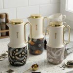 A trio of Feenik 1-liter luxury thermal flasks in cream with gold handles and lids, featuring three different artistic patterns on the base, displayed on a white marble kitchen counter.