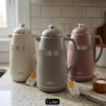 Three 1-liter DayDays thermal flasks in Dusty Rose, Warm Sand, and Stone Grey arranged on a granite kitchen countertop.