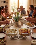 A Paramount Series double stainless steel food warmer on a dining table, featuring two 1.5L glass dishes with lids being lifted to reveal steaming food, supported by a chrome frame with two lit buffet burners underneath.