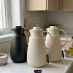 A 1-liter insulated thermal flask with a 3D diamond-patterned matte body, featuring a curved handle and a spout, shown in black and beige color options on a kitchen countertop.