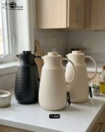 A 1-liter insulated thermal flask with a 3D diamond-patterned matte body, featuring a curved handle and a spout, shown in black and beige color options on a kitchen countertop.