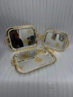 A set of three nesting stainless steel rectangular serving trays with gold rope borders, floral engravings, and ornate gold handles, displayed on a dark wooden dining table with elegant dinnerware.