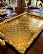 A luxury three-piece set of rectangular gold-plated stainless steel serving trays with intricate floral embossing and ornate vintage handles, displayed on a wooden dining table with elegant place settings.