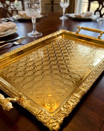 A luxury three-piece set of rectangular gold-plated stainless steel serving trays with intricate floral embossing and ornate vintage handles, displayed on a wooden dining table with elegant place settings.