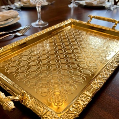 A luxury three-piece set of rectangular gold-plated stainless steel serving trays with intricate floral embossing and ornate vintage handles, displayed on a wooden dining table with elegant place settings.