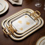 A set of three nesting stainless steel rectangular serving trays with gold rope borders, floral engravings, and ornate gold handles, displayed on a dark wooden dining table with elegant dinnerware.