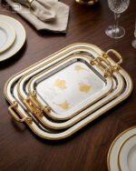 A set of three nesting stainless steel rectangular serving trays with gold rope borders, floral engravings, and ornate gold handles, displayed on a dark wooden dining table with elegant dinnerware.