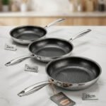 A set of three UAKEEN premium tri-ply stainless steel fry pans with laser-etched honeycomb non-stick interiors, shown in 24cm, 26cm, and 28cm sizes on a marble countertop with an induction-compatible base.