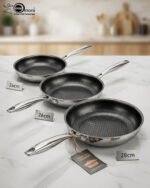 A set of three UAKEEN premium tri-ply stainless steel fry pans with laser-etched honeycomb non-stick interiors, shown in 24cm, 26cm, and 28cm sizes on a marble countertop with an induction-compatible base.