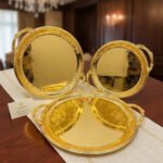 A luxury set of three circular 18K gold-plated stainless steel serving trays featuring intricate floral engravings and ornate handles, displayed on a white runner over a wooden table.