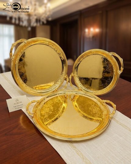 A luxury set of three circular 18K gold-plated stainless steel serving trays featuring intricate floral engravings and ornate handles, displayed on a white runner over a wooden table.