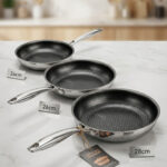 A set of three UAKEEN premium tri-ply stainless steel fry pans with laser-etched honeycomb non-stick interiors, shown in 24cm, 26cm, and 28cm sizes on a marble countertop with an induction-compatible base.