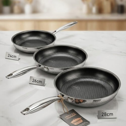 A set of three UAKEEN premium tri-ply stainless steel fry pans with laser-etched honeycomb non-stick interiors, shown in 24cm, 26cm, and 28cm sizes on a marble countertop with an induction-compatible base.