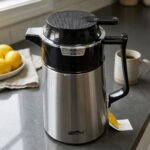 A 1-liter stainless steel vacuum thermal flask with a black insulated lid and ergonomic handle, placed on a kitchen countertop next to lemons and a coffee mug.