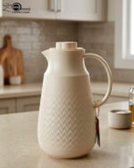 A 1-liter insulated thermal flask with a 3D diamond-patterned matte body, featuring a curved handle and a spout, shown in black and beige color options on a kitchen countertop.