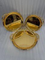 A luxury set of three circular 18K gold-plated stainless steel serving trays featuring intricate floral engravings and ornate handles, displayed on a white runner over a wooden table.