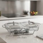 A Paramount Series double stainless steel food warmer on a dining table, featuring two 1.5L glass dishes with lids being lifted to reveal steaming food, supported by a chrome frame with two lit buffet burners underneath.