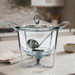 A 4-liter Paramount Series food warmer featuring a clear glass bowl, stainless steel stand, and lid, shown on a dining table with guests and filled with warm lentil soup.