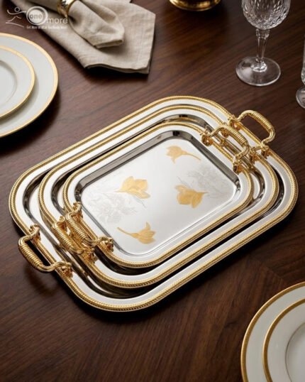 A set of three nesting stainless steel rectangular serving trays with gold rope borders, floral engravings, and ornate gold handles, displayed on a dark wooden dining table with elegant dinnerware.