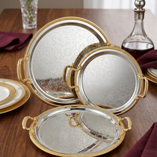 Set of three round silver stainless steel serving trays with floral etched patterns, gold rope borders, and ornate gold handles on a wooden dining table.