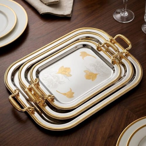 A set of three nesting stainless steel rectangular serving trays with gold rope borders, floral engravings, and ornate gold handles, displayed on a dark wooden dining table with elegant dinnerware.