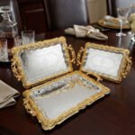 A three-piece set of rectangular stainless steel serving trays featuring polished silver mirror bases with etched ribbon designs and ornate gold-embossed scalloped borders, displayed on a dark wooden dining table.