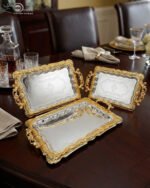 A three-piece set of rectangular stainless steel serving trays featuring polished silver mirror bases with etched ribbon designs and ornate gold-embossed scalloped borders, displayed on a dark wooden dining table.